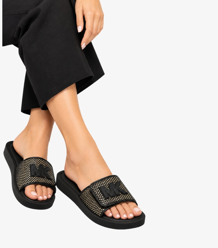 Woman wearing Michael Kors black slide sandals with gold-tone studded straps and raised MK logos