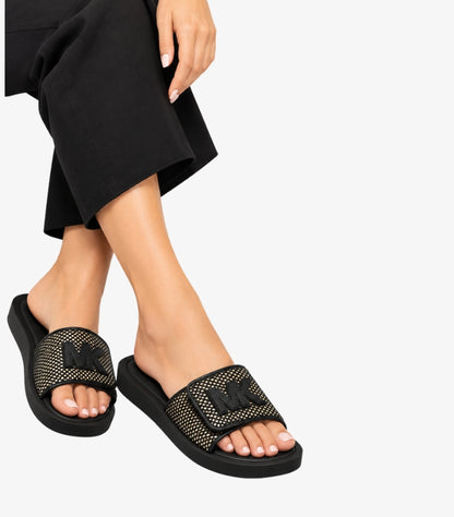 Woman wearing Michael Kors black slide sandals with gold-tone studded straps and raised MK logos