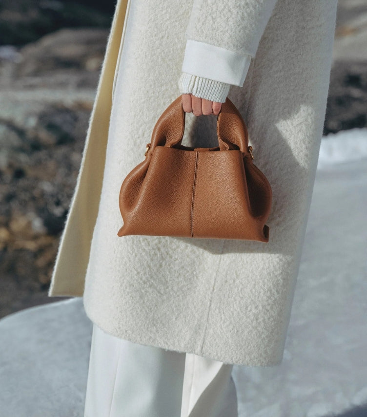 Polène Paris Numéro Nine Mini Sculptural Top Handle Bag in Tan Grained Leather – Angled Front View Carried by Female Model Against White Bouclé Coat Outdoor Setting
