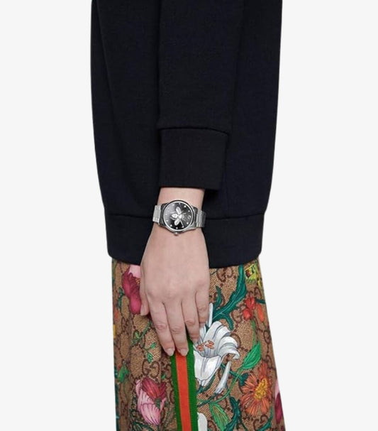 Gucci G-Timeless YA1264136 38mm Black Bee Dial Women's Watch Worn on Wrist – Lifestyle Shot Styled with Black Sweater and Gucci GG Flora Print Skirt Showing Bold Case Size