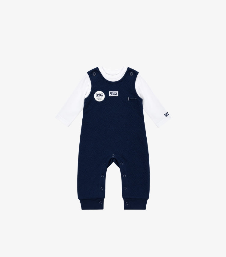 DKNY Jeans baby boys navy blue textured dungaree with logo detailing, buttoned shoulder straps and snap closures, paired with a white long sleeve T-shirt.